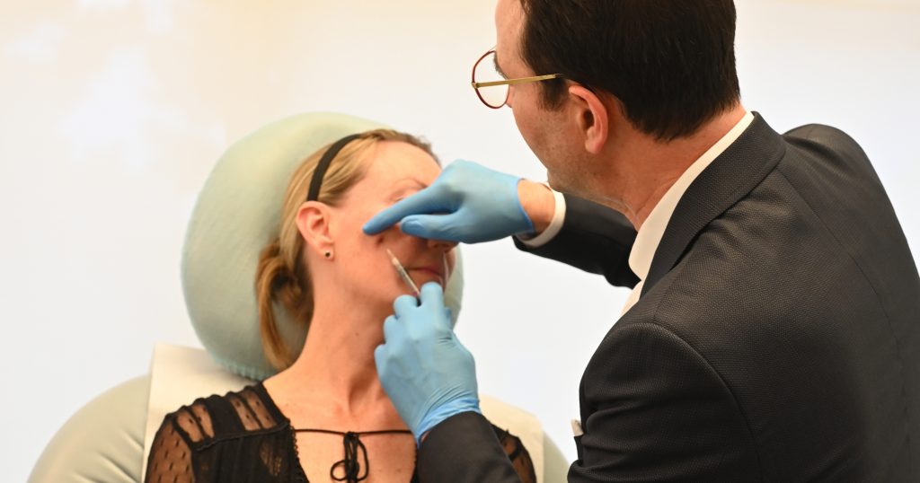 A patient has her eyes closed and is sitting in a chair with her eyes closed while a doctor is resting his hand against her face while getting ready to do an injection. (actual patient)
